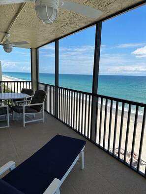 Ein überdachter Balkon mit Blick auf den Strand, ein Tisch mit Stühlen und eine Liege.