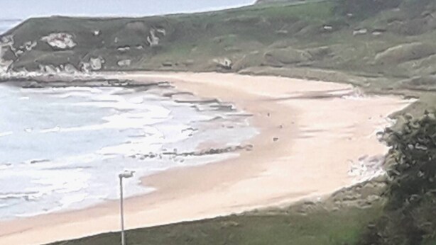 Un paysage côtier comprenant une plage de sable, des collines verdoyantes et une étendue d’eau.