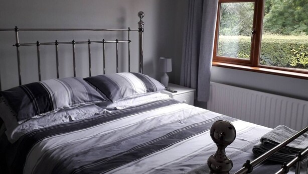 Ein ordentlich bezogenes Bett mit Streifenmuster, ein Nachttisch mit Lampe und ein Fenster mit Blick auf Grünflächen.