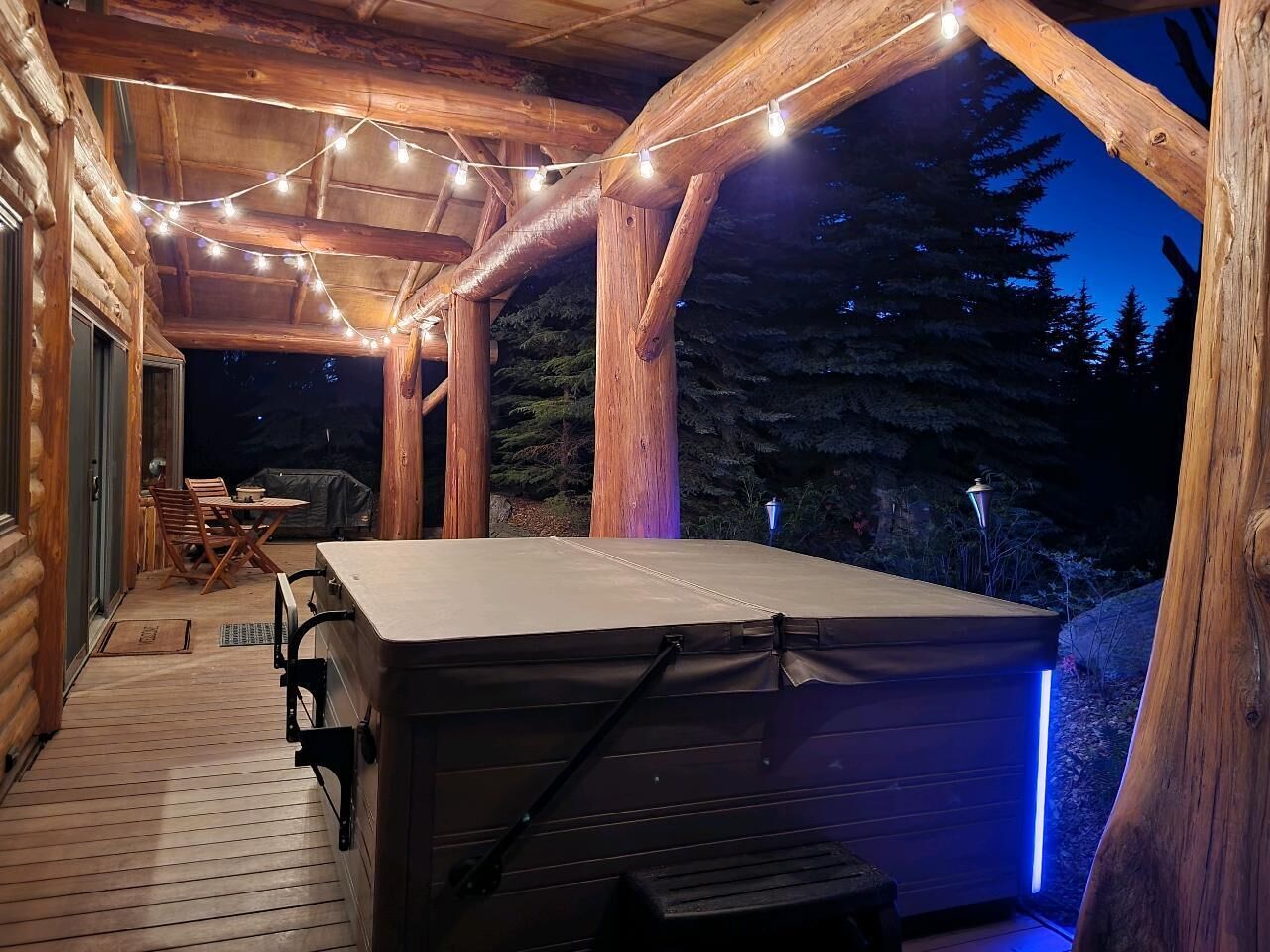 Un bain à remous avec une terrasse en bois et des guirlandes lumineuses, entouré d’une structure de cabane en bois.