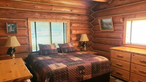 A log cabin bedroom with a bed, two nightstands, a dresser, and two windows with blinds.