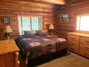 A log cabin bedroom with a bed, two nightstands, a dresser, and two windows with blinds.