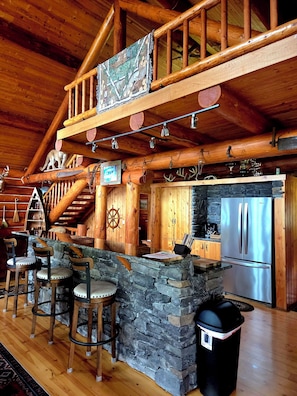 A rustic wooden interior with a stone bar, wooden stools, a stainless steel refrigerator, and a wooden staircase.