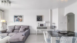 A modern living room with a grey sectional sofa, a dining table with chairs, a small white side table, and a wall-mounted picture frame.