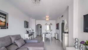 A modern living room with a grey sofa, a dining table with chairs, a white cabinet, and a decorative mirror.