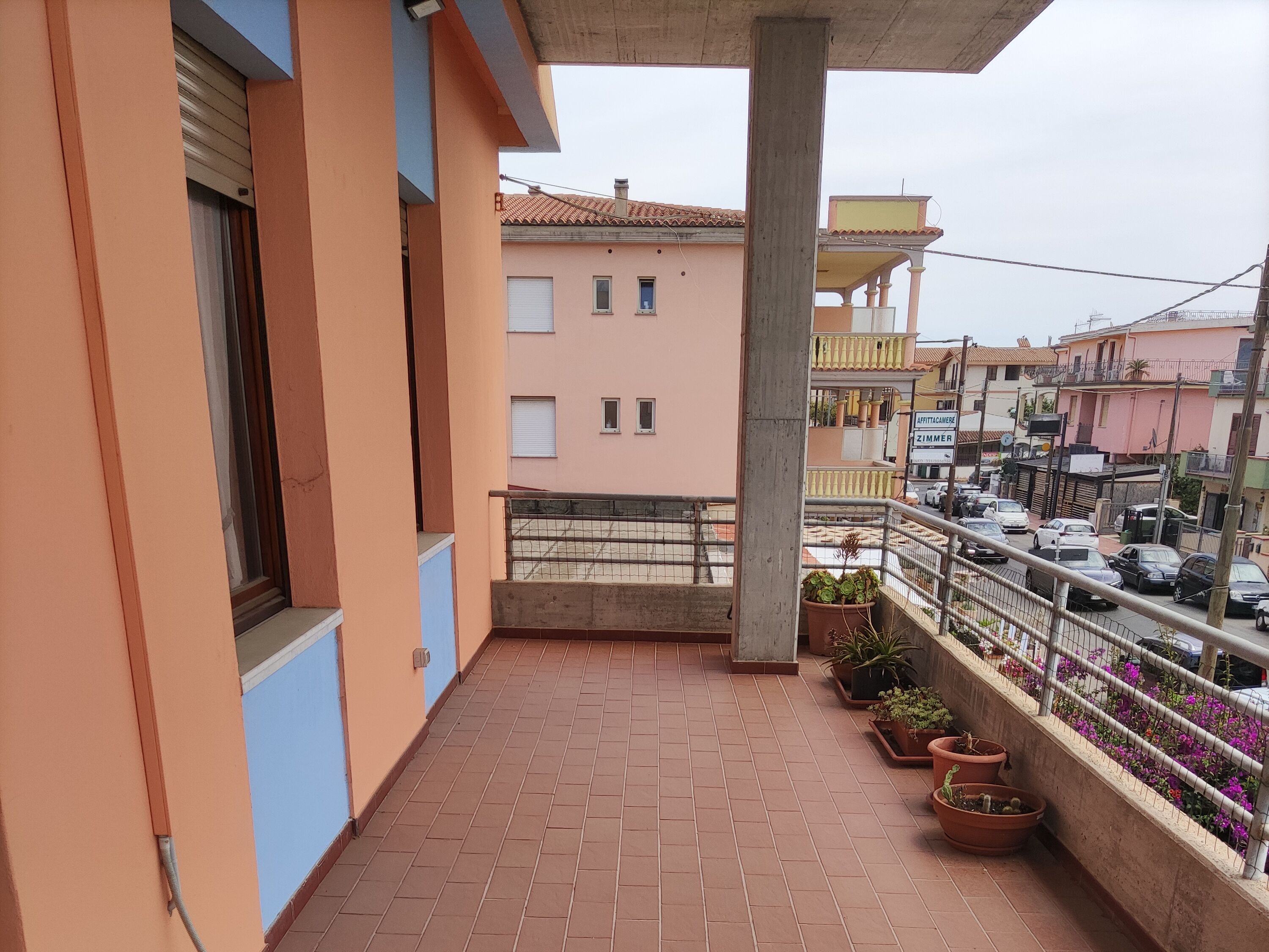 Ein Balkon mit Fliesenboden, Blumentöpfen und Blick auf eine Straße mit parkenden Autos und Gebäuden.