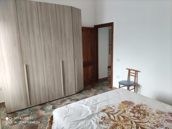 Ein Schlafzimmer mit Kleiderschrank, einem Bett mit floralem Überwurf, einem Sessel und einem Fliesenboden.