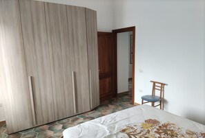 A bedroom with a wardrobe, a bed with a floral bedspread, a chair, and a tiled floor. - Renovated vacation house with terrace, only 270 meters from the beach. (La Caletta)