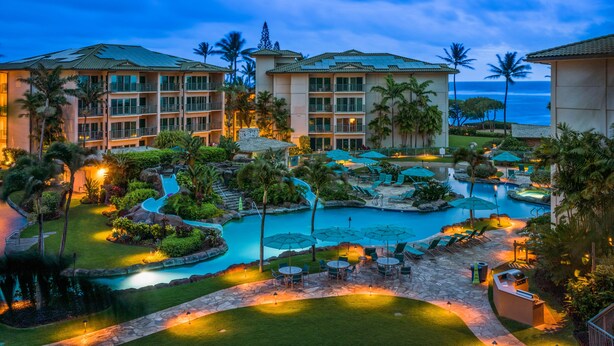 Un resort con varias edificaciones, un área de piscina central con sillones y sombrillas, y un jardín paisajístico circundante.