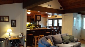 A cozy living room with a blue sofa, wooden bar stools, and a kitchen area with wooden cabinets and a bar counter.