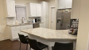 Una cocina con armarios blancos, encimera de granito, refrigerador, microondas y dos taburetes de bar.