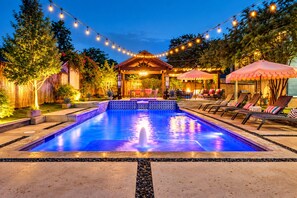 A swimming pool with a tiled edge, surrounded by lounge chairs and umbrellas, under string lights at dusk. - Backyard Resort: Heated Pool, Spa & Putting Green (Fort Worth)