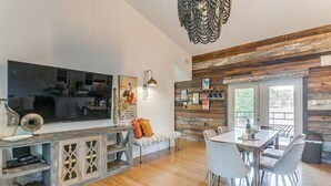 A modern living room with a large flat-screen TV, a wooden dining table, and a cozy seating area. - Backyard Resort: Heated Pool, Spa & Putting Green (Fort Worth)