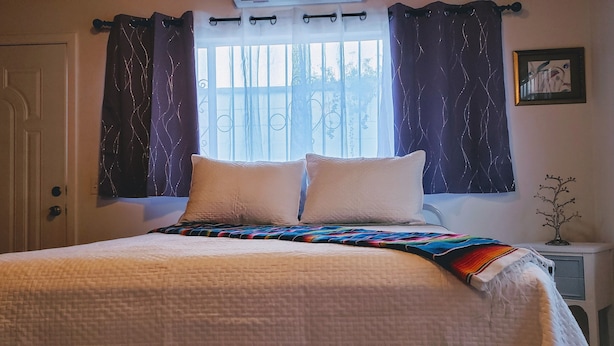 A neatly made bed with a colorful blanket, a window with purple curtains, and a small nightstand with a plant. - Central, Modern, Fast Wi-fi. Beach, food, nightlife & market access w/in 3 km (Ensenada)