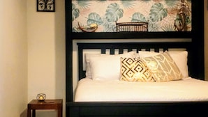 A bed with a black headboard, white bedding, and patterned pillows. Above the bed, there is a shelf with decorative items and a wall with a leaf-patterned wallpaper. - Pura Vida Casita Retreat: Temp-control heat+more modern conveniences for comfort (Ensenada)