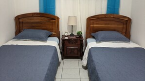 A room with two wooden beds, each with a blue bedspread and pillow, a nightstand with a lamp, a small clock, and a framed picture on the wall. - Entire Comfy apartment for your,  5 min SJO airport (Alajuela)