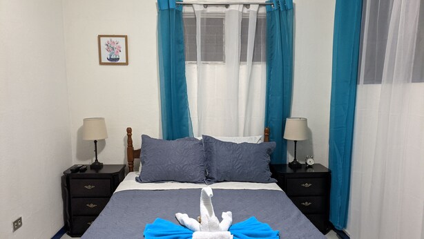A neatly made bed with a towel and a plush toy in the center, flanked by two nightstands with lamps and a framed picture on the wall. - Entire Comfy apartment for your,  5 min SJO airport (Alajuela)