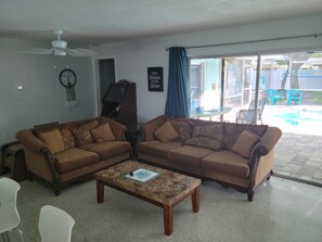 A living room with two brown sofas, a coffee table, a ceiling fan, and a large sliding glass door leading to an outdoor area with a pool. - Salt Pool blocks to beach Gas heated Pool! East side of A1A 3 blocks to beach (Cape Canaveral)