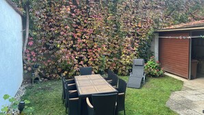 Un patio con mesa y sillas, rodeado por un jardín con diversas plantas y un arbusto grande de hojas multicolores.