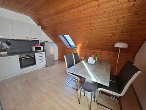 A modern kitchen with white cabinets, a stainless steel refrigerator, and a microwave. There is a dining table with chairs and a vase with flowers. A skylight is visible above the kitchen.