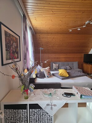 A cozy room with a wooden ceiling, a bed with grey bedding and a yellow pillow, a bedside table with a glass vase and a book, and a framed picture on the wall.