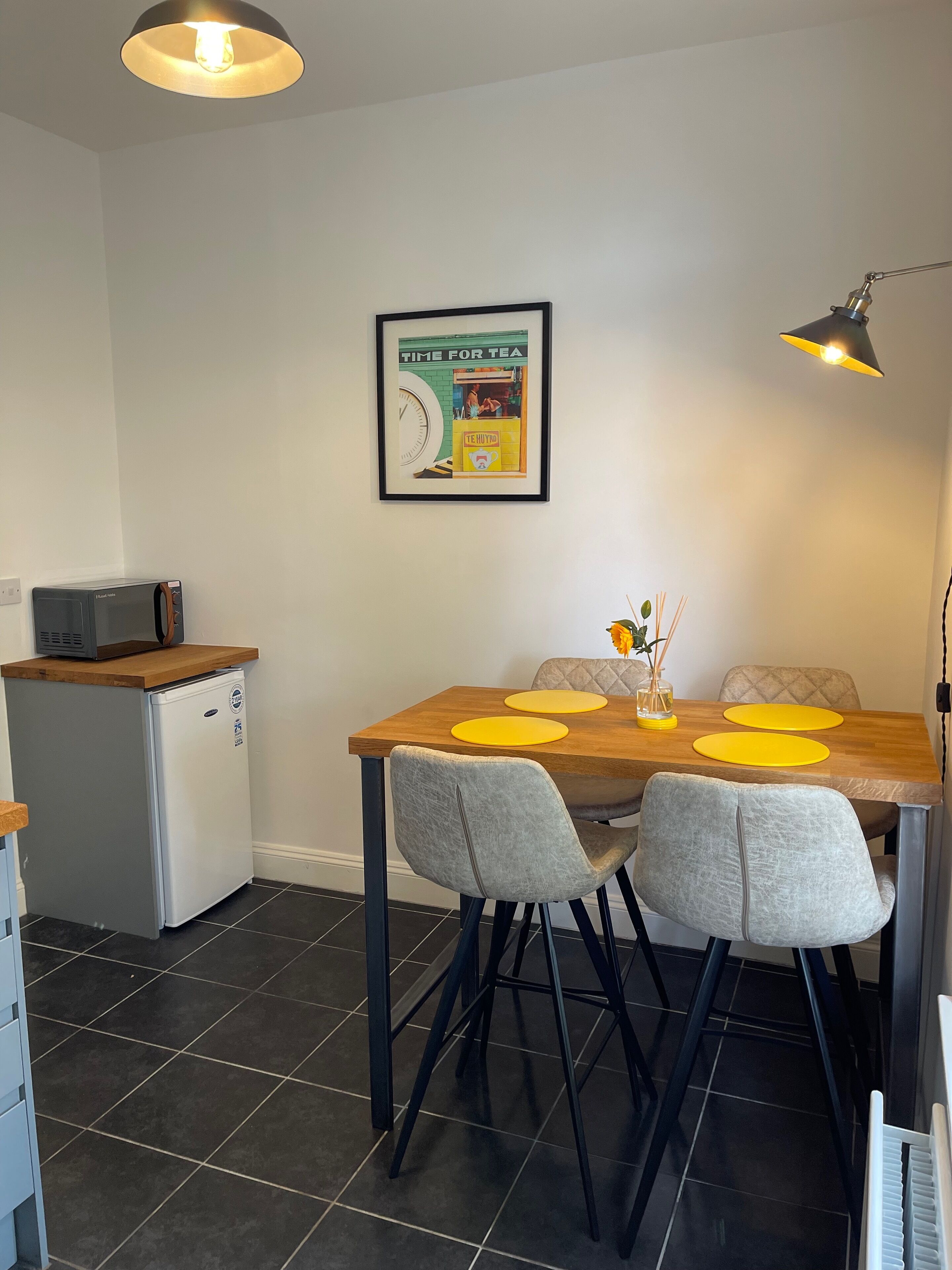 Una cucina compatta con zona pranzo, che include un frigorifero, un forno a microonde e un poster incorniciato appeso al muro.