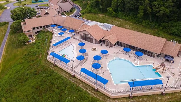 Luchtbeeld van een resort met een groot zwembad, buitenzitplaatsen en een gebouw met een schuin dak.