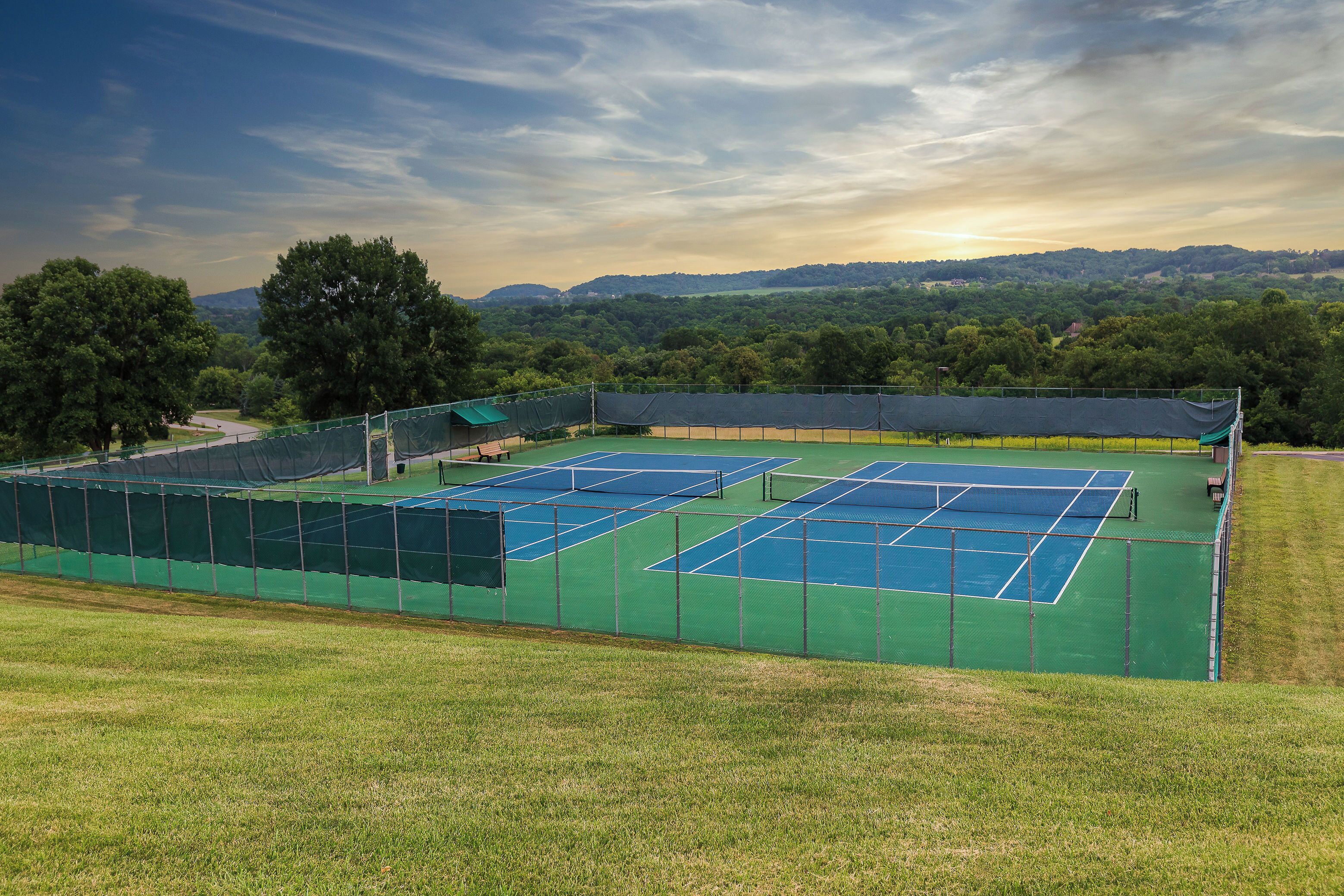 Canchas de tenis múltiples, rodeadas por una cerca verde, con vistas a un paisaje montañoso al fondo.