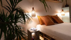 Un dormitorio con una cama, dos lámparas de noche, un estante con una planta y una mesita con libros.