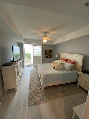 Relax in a spacious bedroom with a large bed, ceiling fan, and TV, offering a view of lush greenery through large windows.