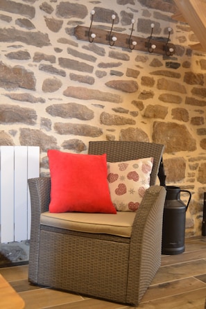 Une chaise en osier avec un coussin rouge et un oreiller à motifs, placée contre un mur de pierre, au-dessus duquel se trouve un porte-manteau en bois.