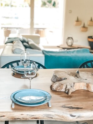 Una mesa de comedor con platos azules y copas de vino, un centro de mesa de madera y una zona de estar acogedora al fondo.