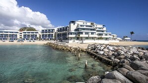 Un hotel frente a la playa con varios pisos, un rompeolas rocoso y aguas azules cristalinas.
