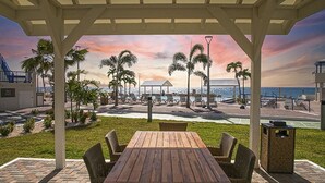Un patio cubierto con una mesa y sillas de madera, con vista a una playa con palmeras y un atardecer.