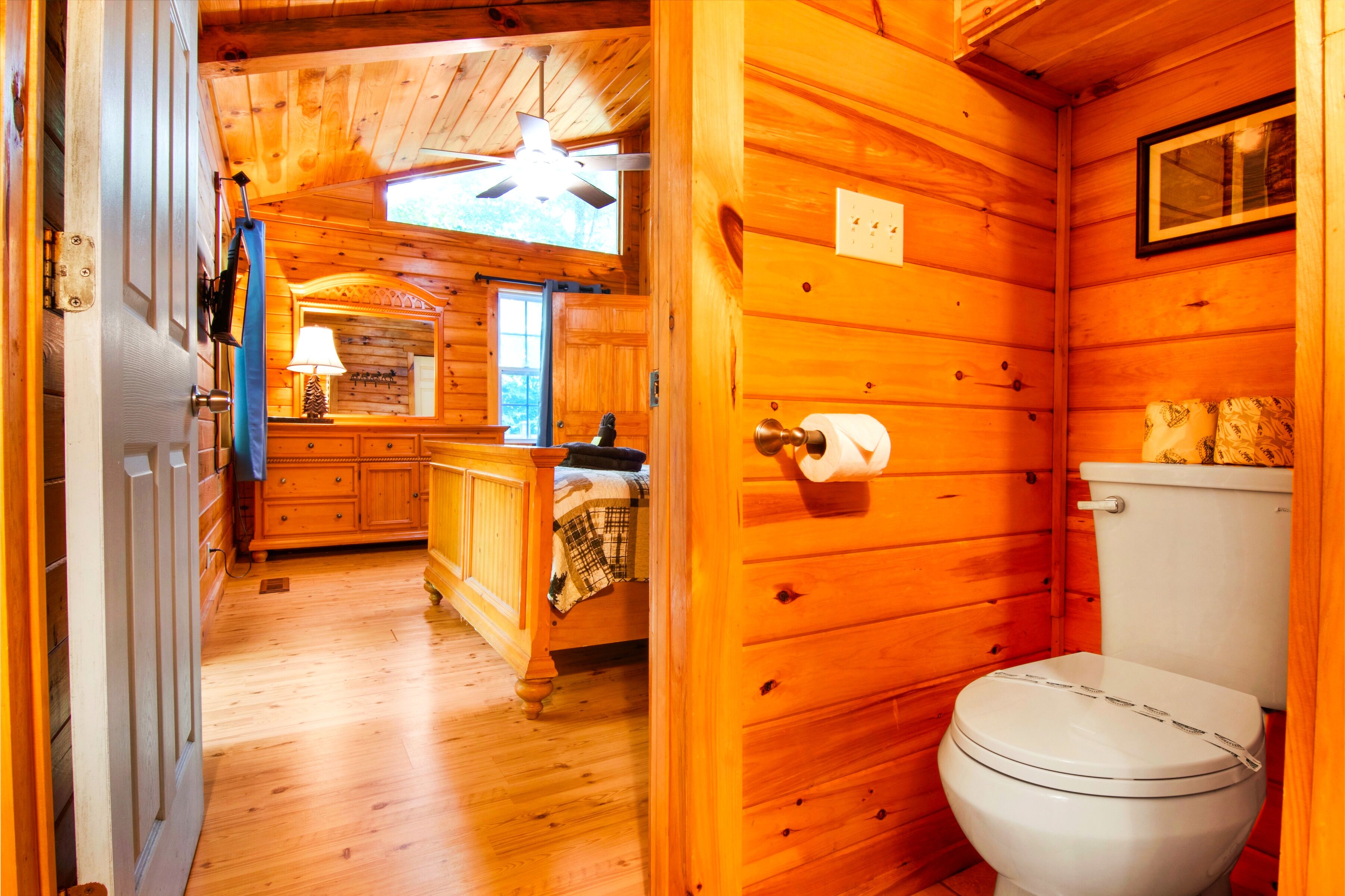 Ein Holzhaus-Interieur mit Holzboden, einer weißen Toilette, einem Holzschrank, einem Deckenventilator und einem Fenster.
