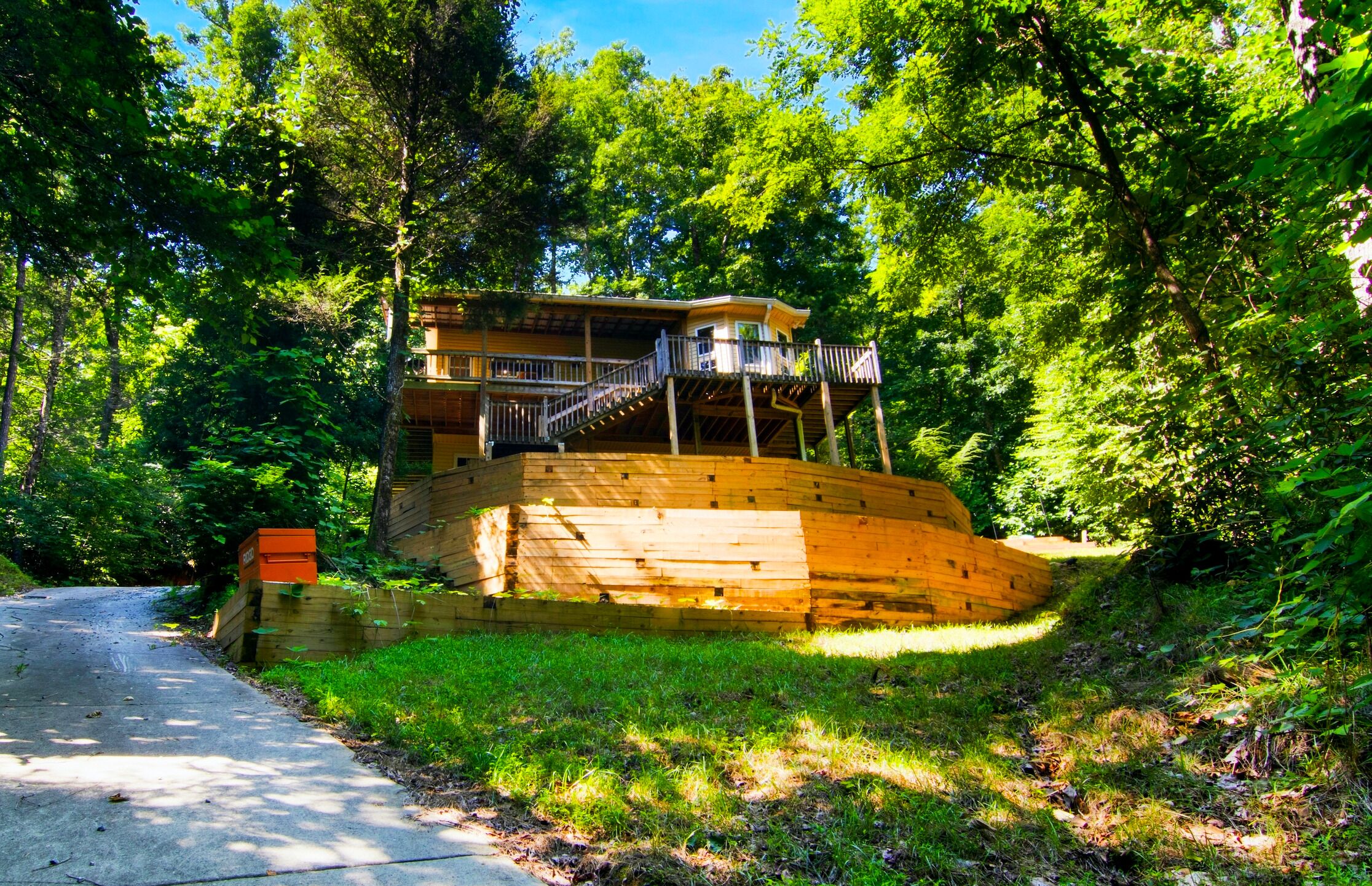 A wooden cabin with a balcony, surrounded by lush greenery and a dirt path.