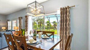 A dining area with a table set for four, surrounded by wooden chairs, a chandelier, and a view of a swimming pool and greenery. - Bay Lilly Family Villa Close to Parks sleeps 8 (Kissimmee)