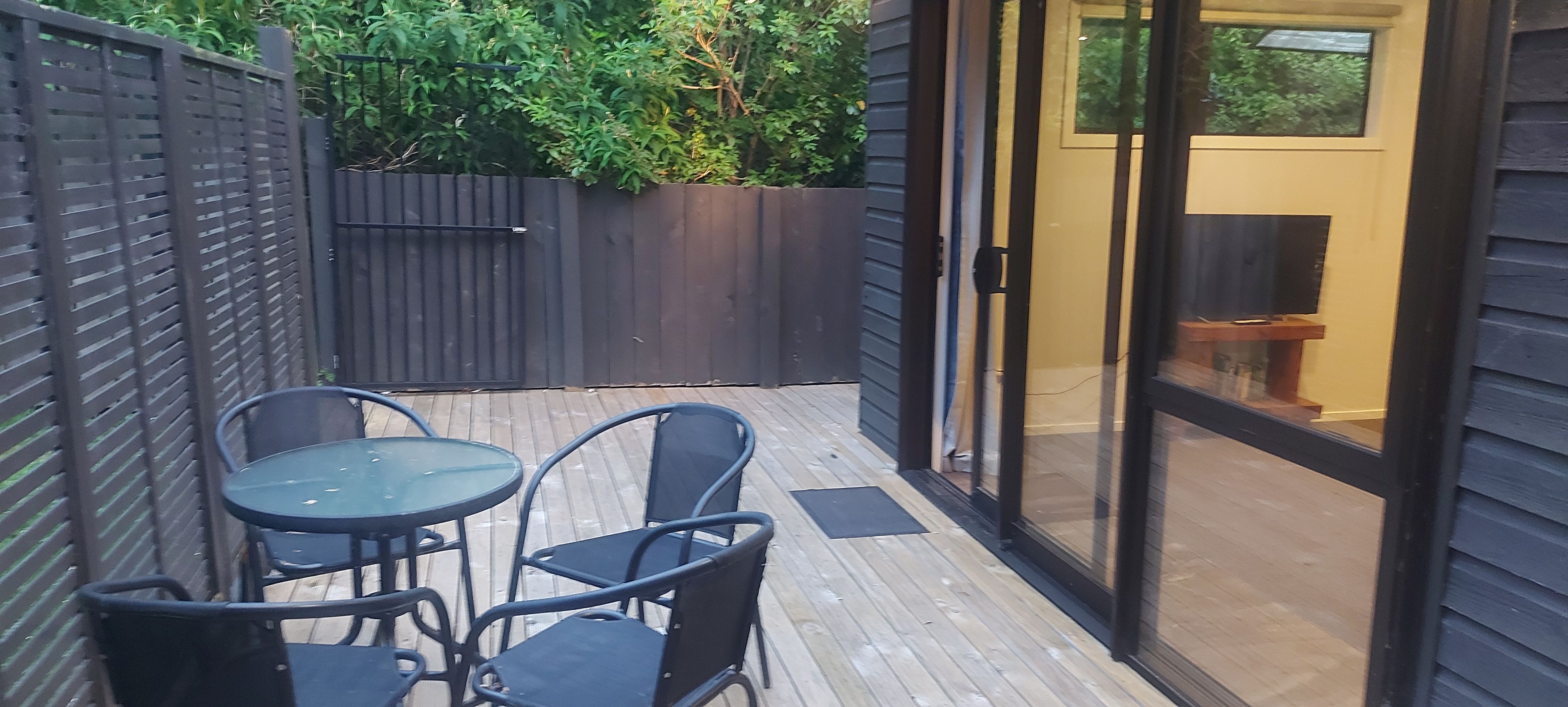 A patio with a table and chairs, a wooden deck, a glass door leading inside, and a dark fence.