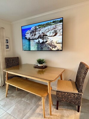 A dining area with a wooden table, two patterned chairs, a bench, and a flat-screen TV mounted on the wall displaying a nature scene.