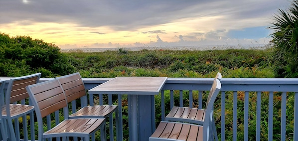 A wooden deck with two chairs and a table, overlooking a grassy area with a view of the ocean.