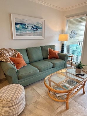 A living room with a green sofa, two orange pillows, a glass coffee table, a wicker chair, a framed picture on the wall, and a window with blinds.