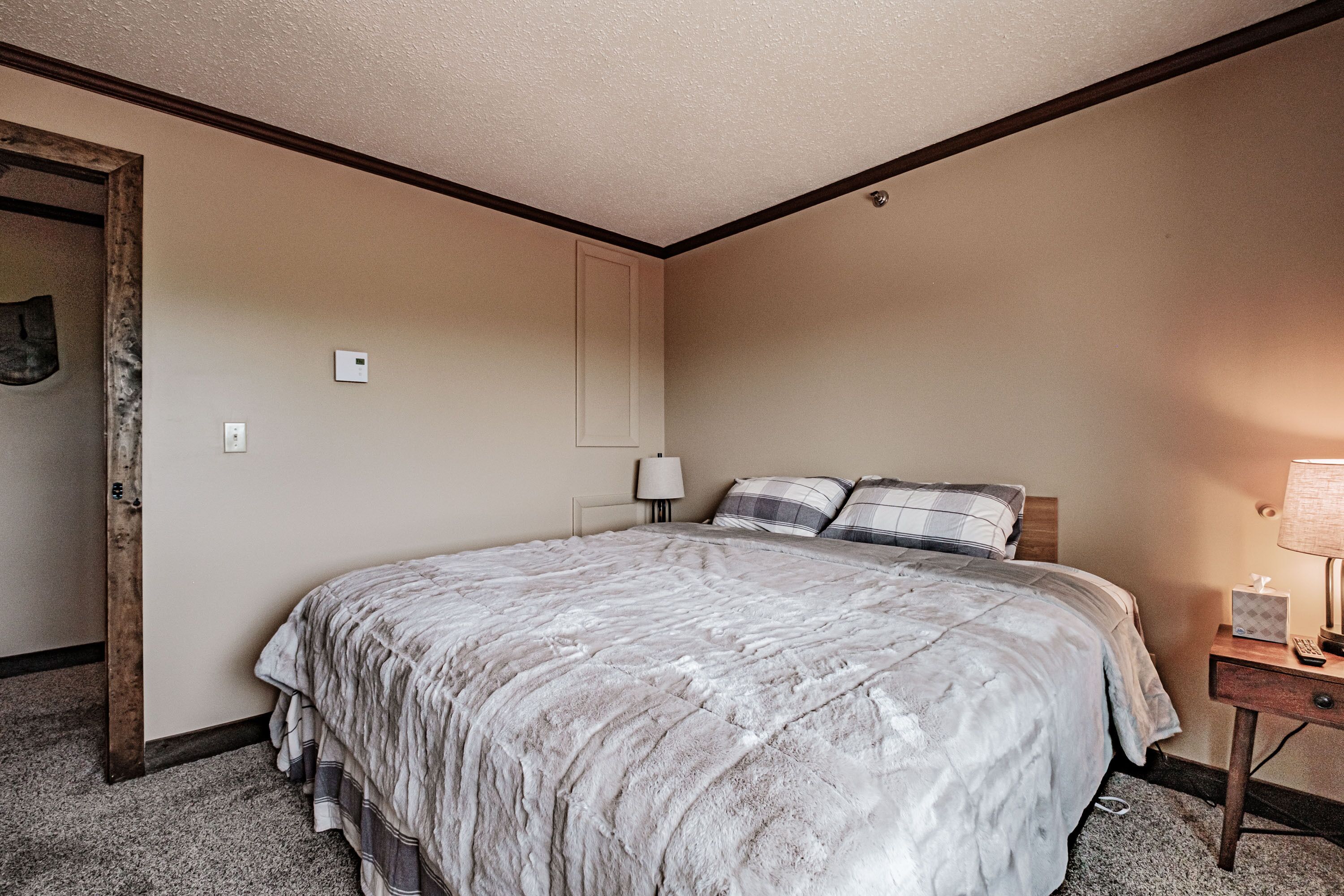 Una habitación de hotel con una cama grande, una mesita de noche con lámpara, y una puerta que da a otra habitación.