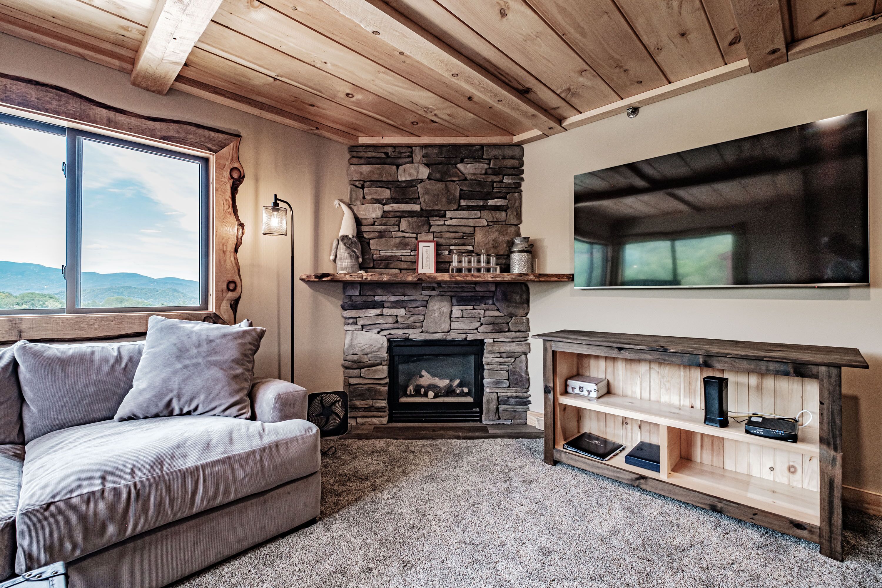 A cozy living room with a stone fireplace, a large window, and a wooden ceiling.