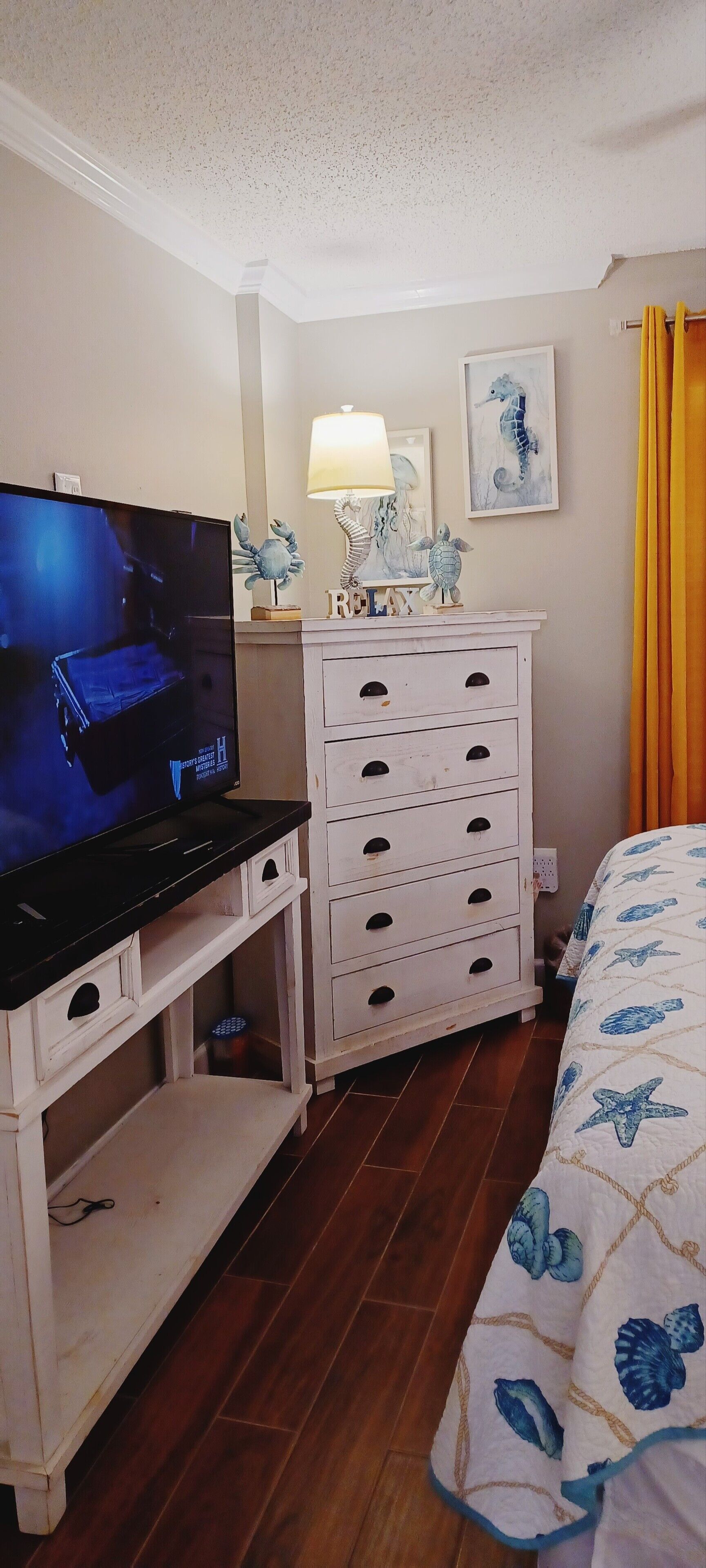 A hotel room with a television on a stand, a dresser with drawers, a bed with a starfish-patterned bedspread, and a framed seahorse artwork on the wall.