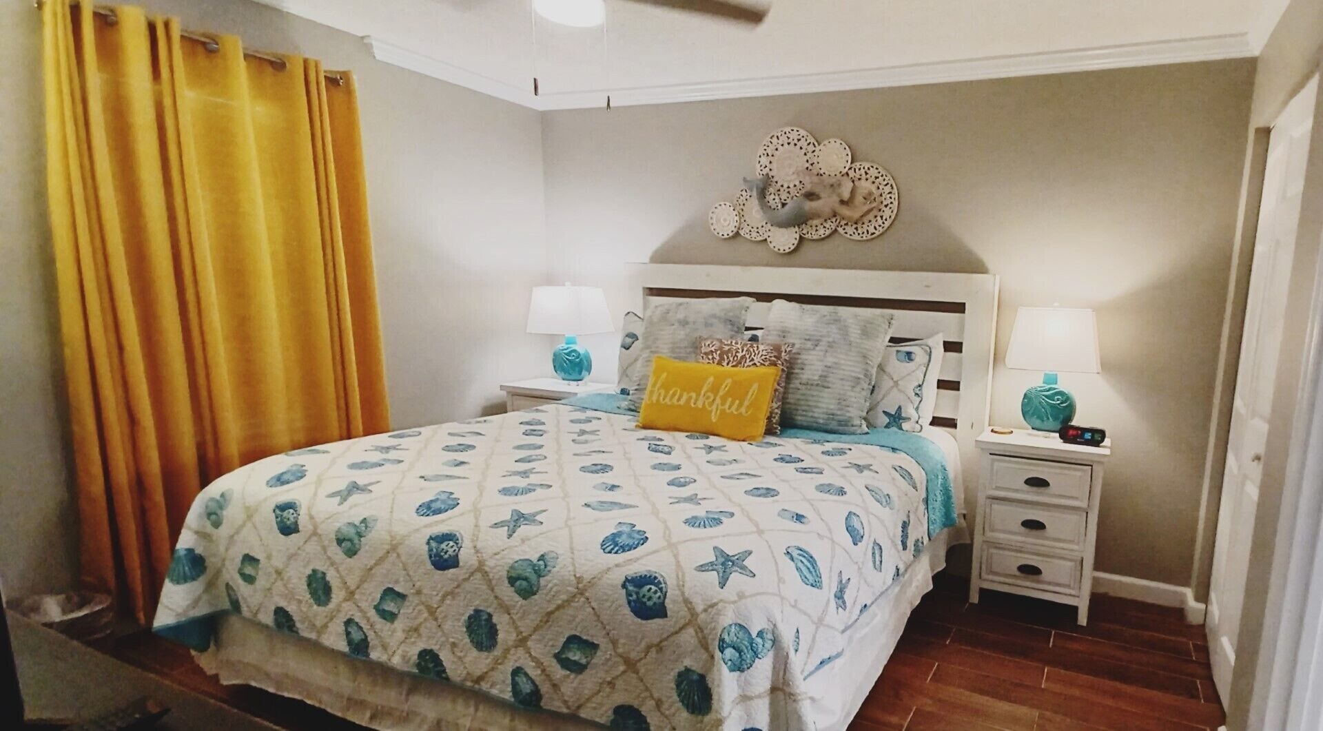 A bedroom with a bed, two bedside tables, a nightstand, a wall decoration, and a window with yellow curtains.
