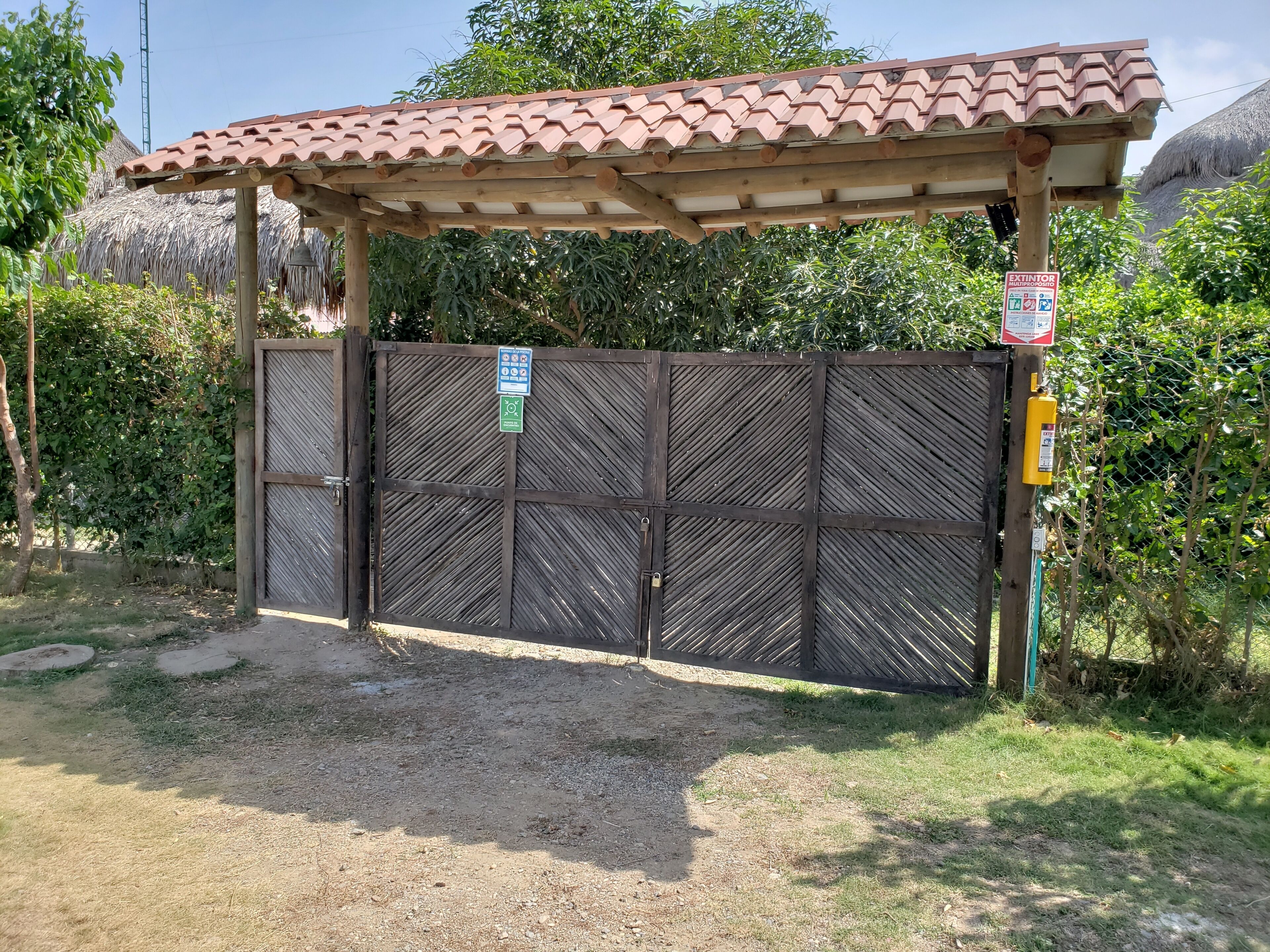 A gated entrance with a wooden pergola overhead, a sign indicating an extinctor, and a thatched roof structure in the background.