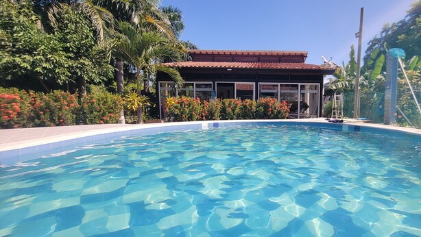 A clear swimming pool with a tiled edge, surrounded by lush greenery and a building with a tiled roof in the background. - Indigo Palomino Beautiful House (Palomino)
