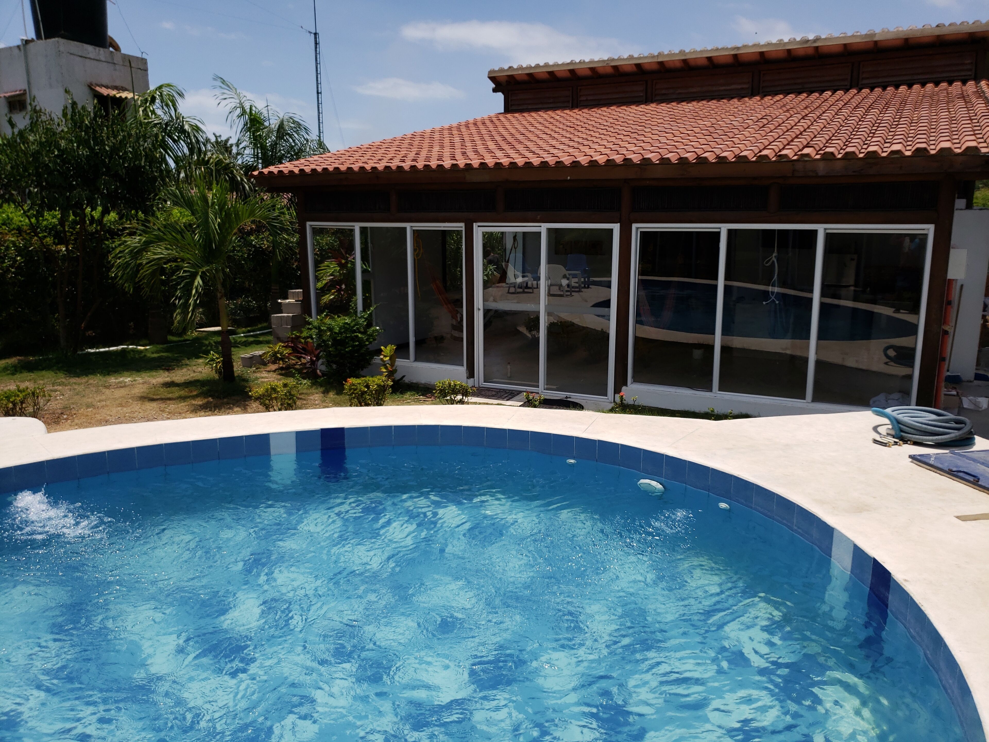A swimming pool with a tiled edge, a covered patio area with a tiled roof, and a view of a residential building with large windows.