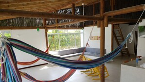 A hammock suspended from a wooden ceiling in a room with a white tiled floor, a window with a view of greenery, and a yellow rocking chair. - Indigo Palomino Beautiful House (Palomino)