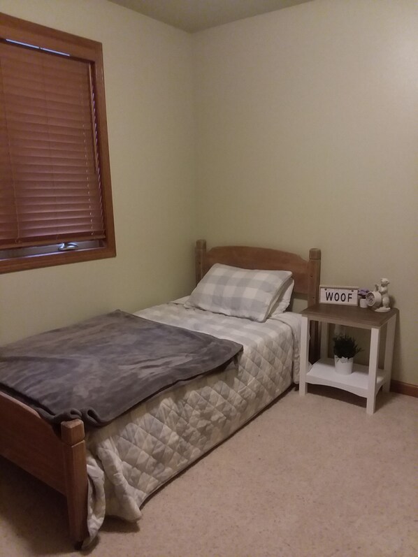 A single bed with a quilted bedspread, a bedside table with a small plant, and a sign that reads "WOOF".